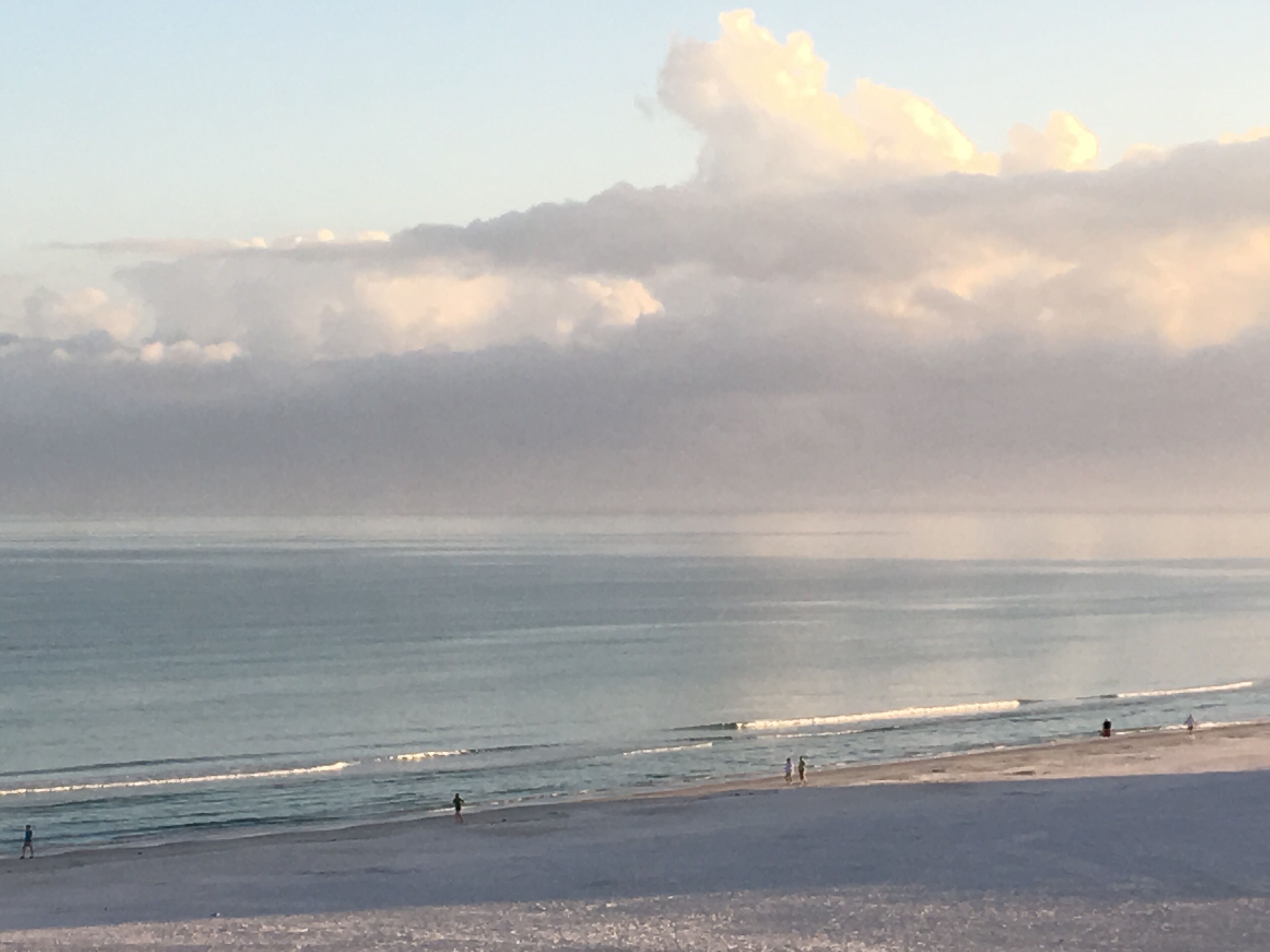 A beach with a clear sky and clouds, calm sea, and a few people walking on the shore.