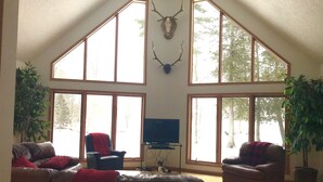 Un salon spacieux avec un plafond haut, de grandes fenêtres et une tête de cerf accrochée au mur. On y trouve deux canapés en cuir, un téléviseur et une table basse.