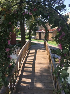 A wooden bridge with a metal railing, surrounded by greenery and a stone building in the background. - RistHotel Pianura Inn (Busnago)