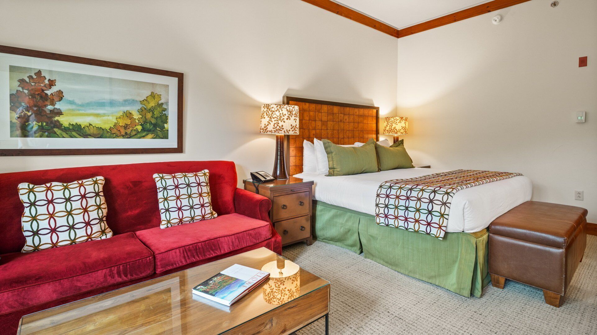 Een hotelkamer met een rode bank, een houten nachtkastje, een bed met een groene plaid en een lijst met een landschap op de muur.