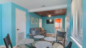 A dining area with a glass table surrounded by chairs, a sofa, and a television in the background.