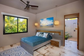 Un dormitorio con cama, mesitas de noche, ventilador de techo, un ventanal con vistas a la vegetación y un cuadro en la pared.