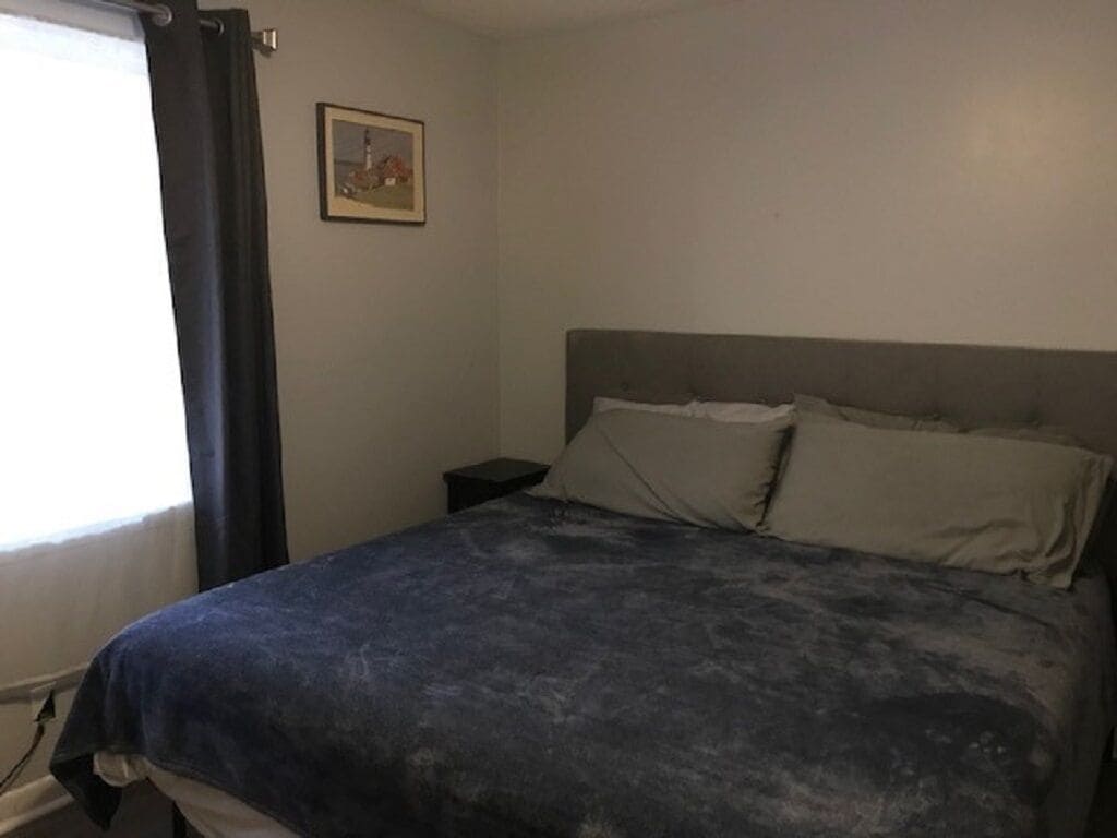 A bedroom with a bed, a nightstand, a picture on the wall, and a window with curtains.