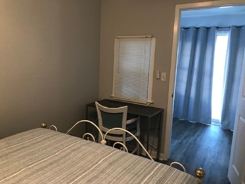 Ein Schlafzimmer mit einem Bett, einem Schreibtisch und einem Stuhl. Es gibt ein Fenster mit Jalousien, eine zur Außenwelt führende Tür und einen Vorhang.