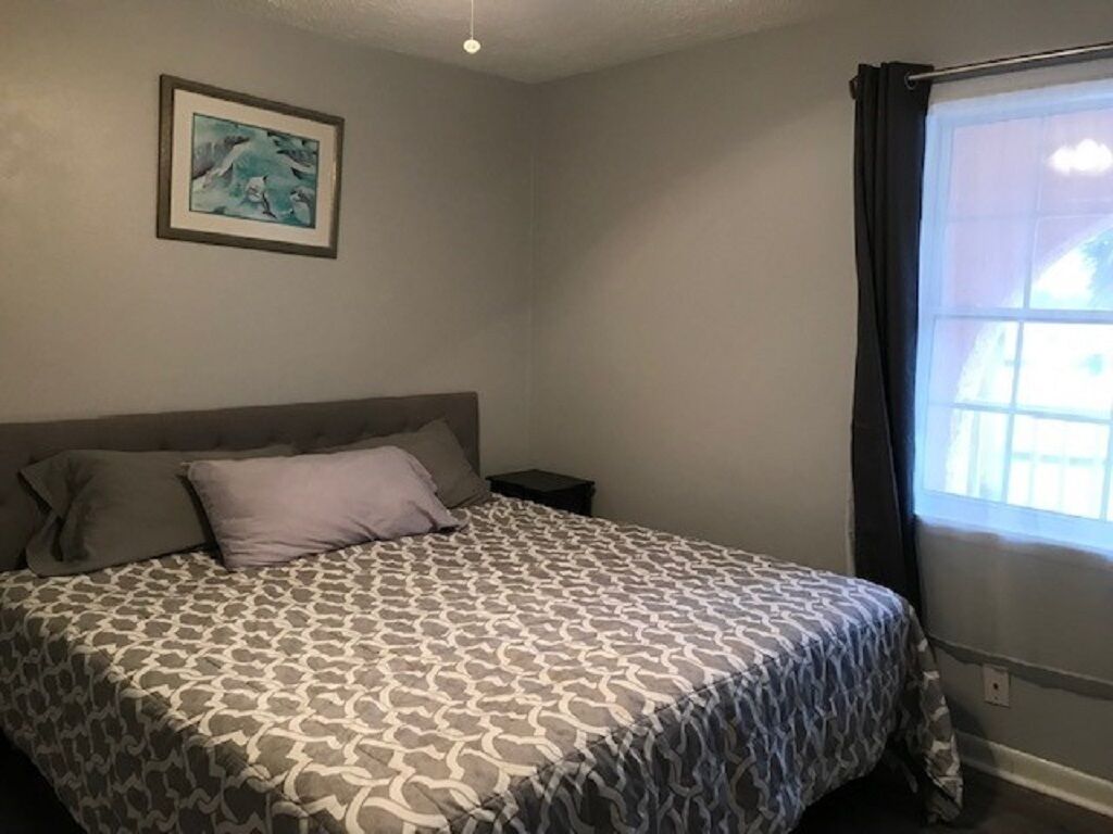Un dormitorio con cama, mesita de noche, un cuadro enmarcado en la pared y una ventana con cortinas.