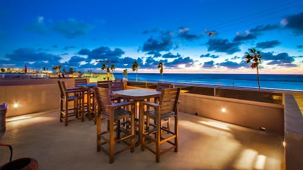 A terrace with wooden tables and chairs, overlooking a beach at sunset.