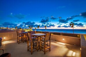A terrace with wooden tables and chairs, overlooking a beach at sunset. - Surf & Sand | 3BR Mission Beach Steps to Shore (San Diego)