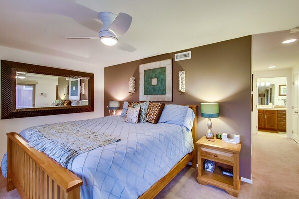A bedroom with a large bed, a bedside table with a lamp, a ceiling fan, and a wall-mounted mirror.