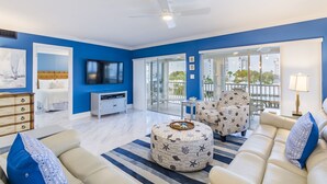 Una sala de estar moderna con una pared azul de acento, un ventilador de techo, un televisor de pantalla plana, un sofá, otomanas y un comedor con mesa y sillas.