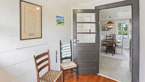 Una habitación con una silla de madera, una puerta con un letrero, un mapa enmarcado y vista a otra habitación con una mesa y sillas.