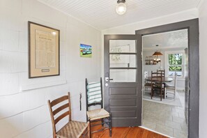 A room with a wooden chair, a door with a sign, a framed map, and a view into another room with a table and chairs.