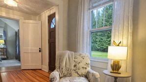 Una habitación con piso de madera, un sillón floral, una pequeña mesa con una lámpara y vistas a zonas verdes a través de una ventana.