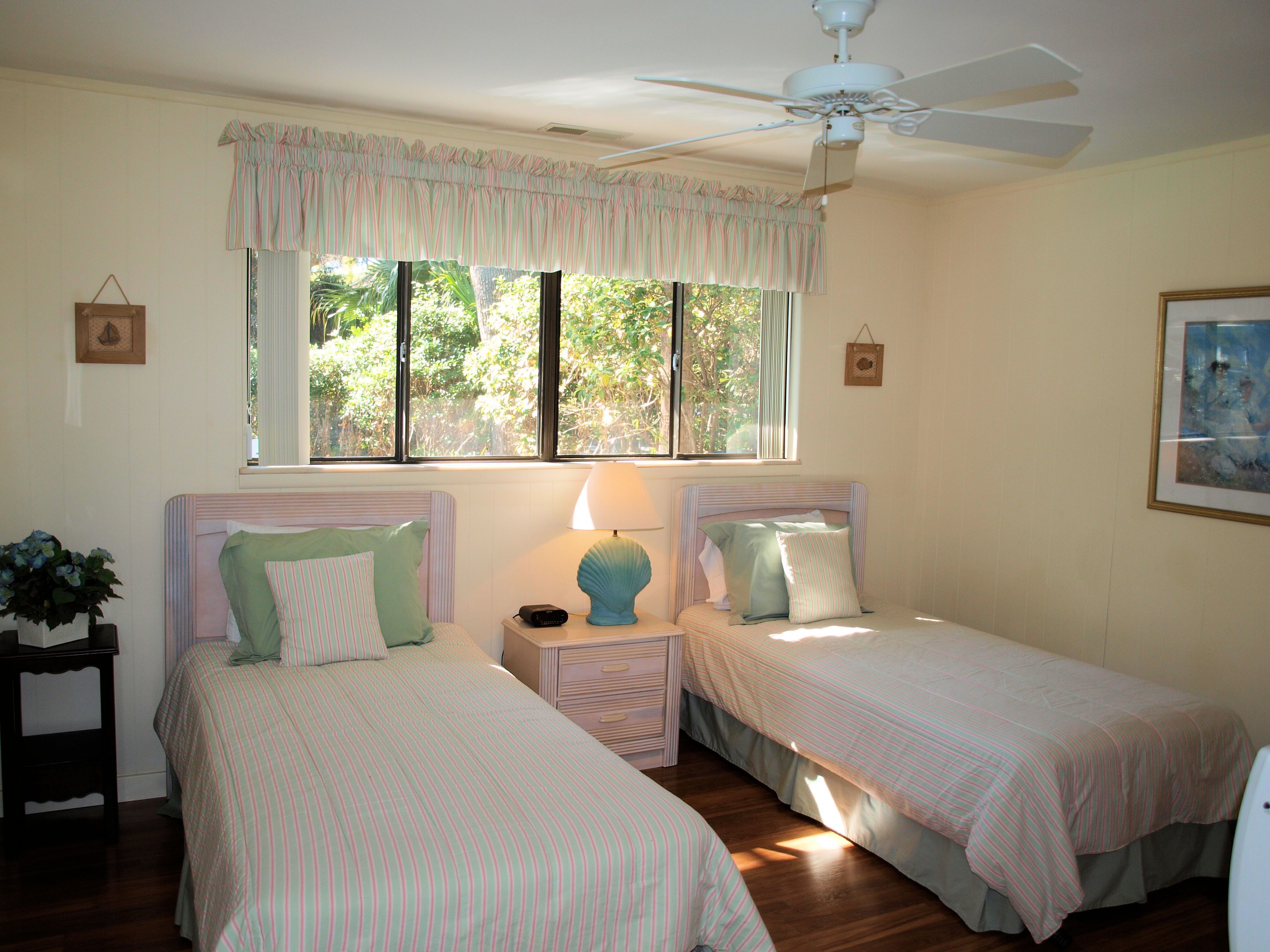 A bedroom with two single beds, a ceiling fan, a window with curtains, and a bedside table with a lamp.