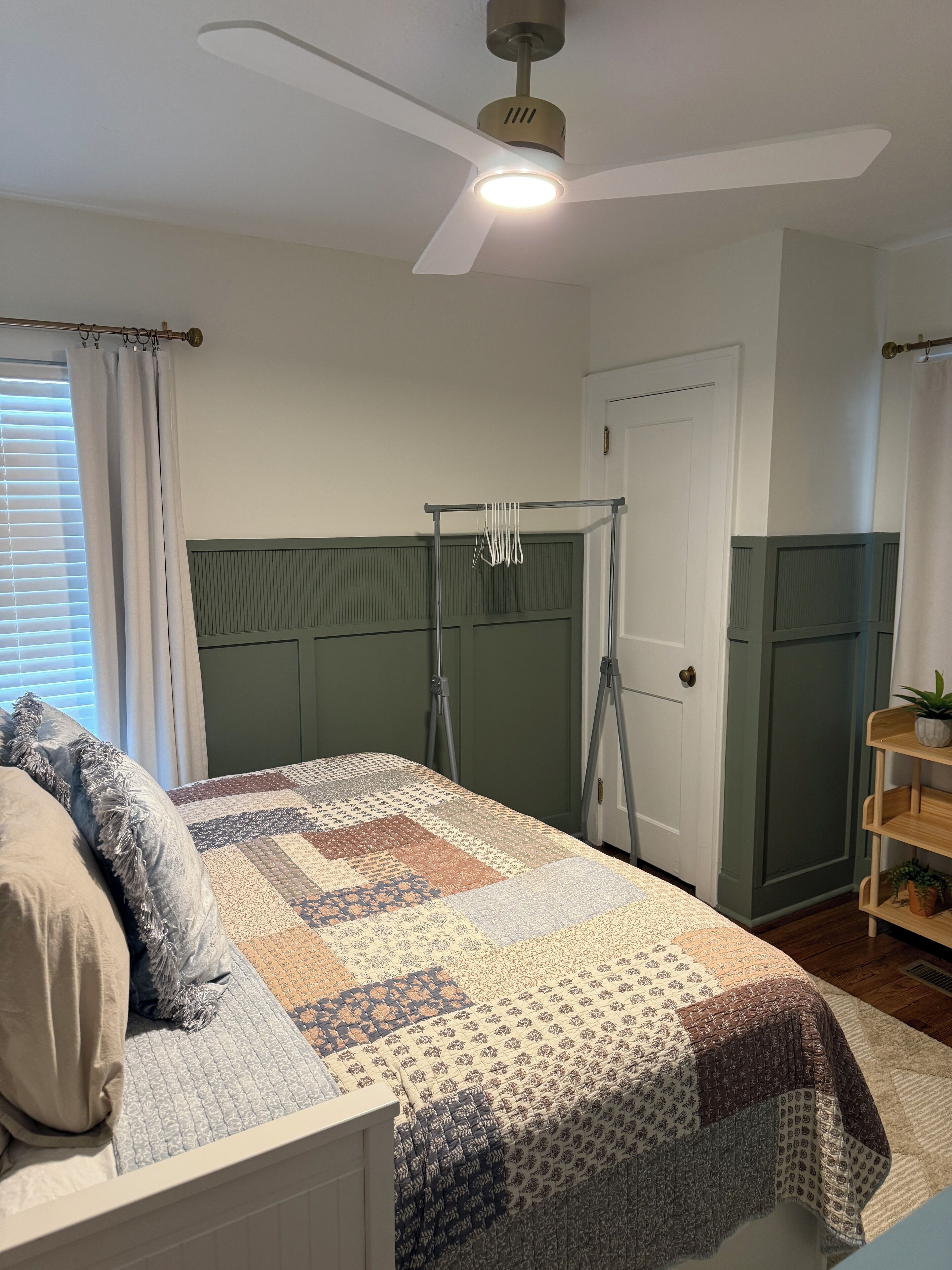 A bedroom with a bed, a ceiling fan, a window with curtains, a door, and a small shelf with a plant.
