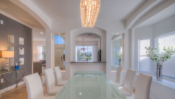 A spacious dining area with a glass table and white chairs, a chandelier, and a view of the outdoors through large windows. - Lakeside house, huge south facing pool with spa,  lake view, Disney at 8 miles (Kissimmee)