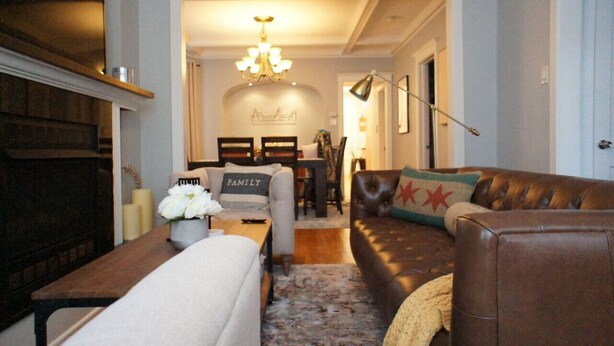 Ein Wohnzimmer mit einer braunen Ledersofa, einem weißen Sessel und einem Holztisch. Eine Kristallleuchter hängt von der Decke und ein gerahmtes Bild ist an der Wand.
