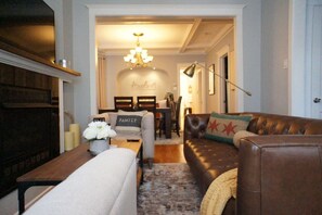 A living room with a brown leather sofa, a white armchair, and a wooden coffee table. There is a chandelier hanging from the ceiling and a framed picture on the wall.