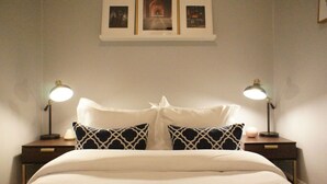Ein ordentlich bezogenes Bett mit weißer Bettwäsche und zwei Zierkissen, begleitet von zwei Nachttischen mit Lampen, in einem schlichten Zimmer mit weißer Wand und einem gerahmten Bild über dem Bett.