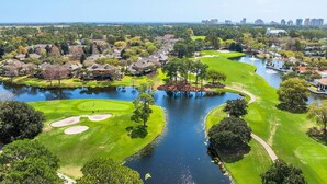 Campo de golf con obstáculos de agua, rodeado de edificios residenciales y un horizonte urbano a lo lejos.