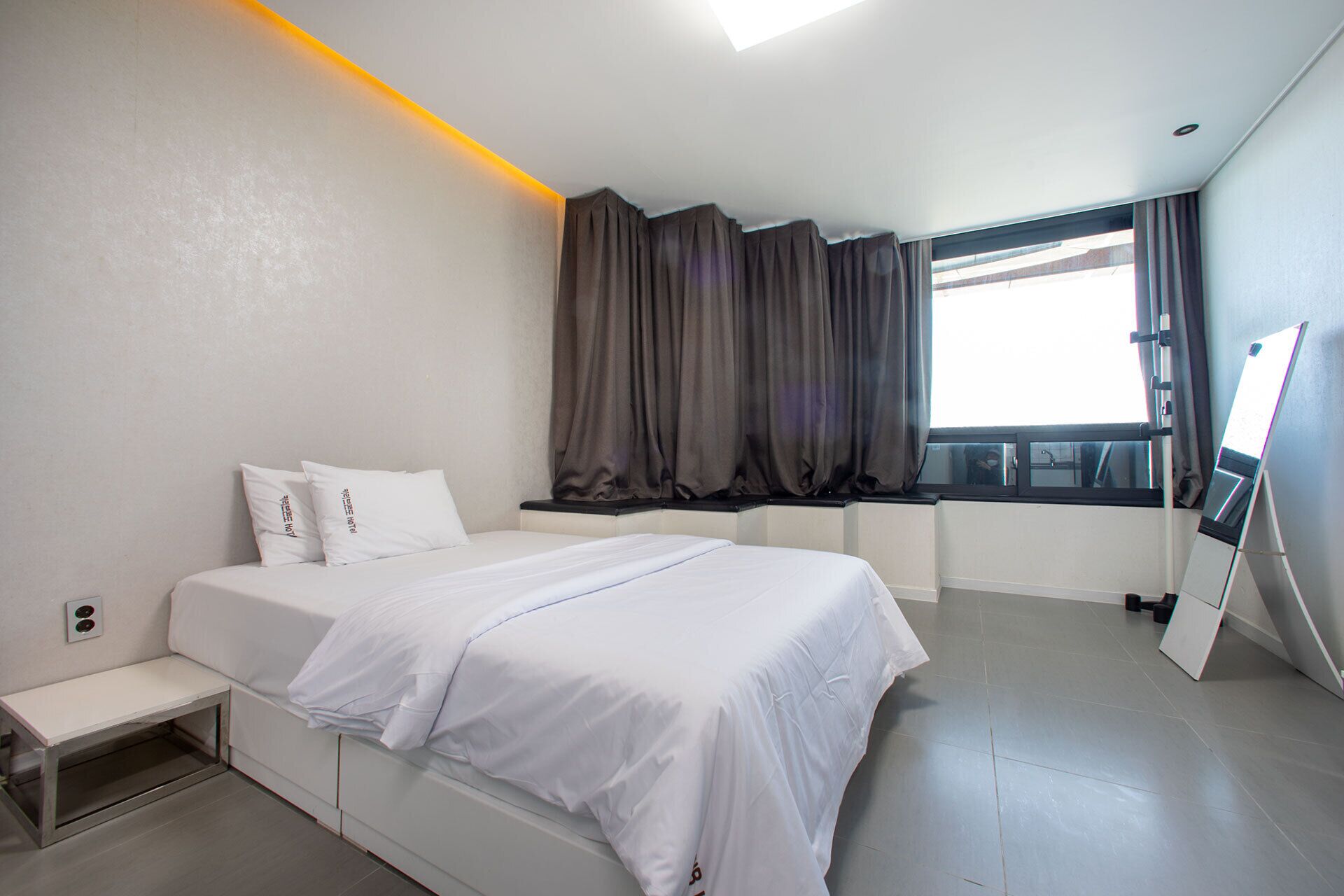 Una habitación de hotel moderna con cama, una mesita de noche pequeña, una ventana con cortinas y un espejo grande.