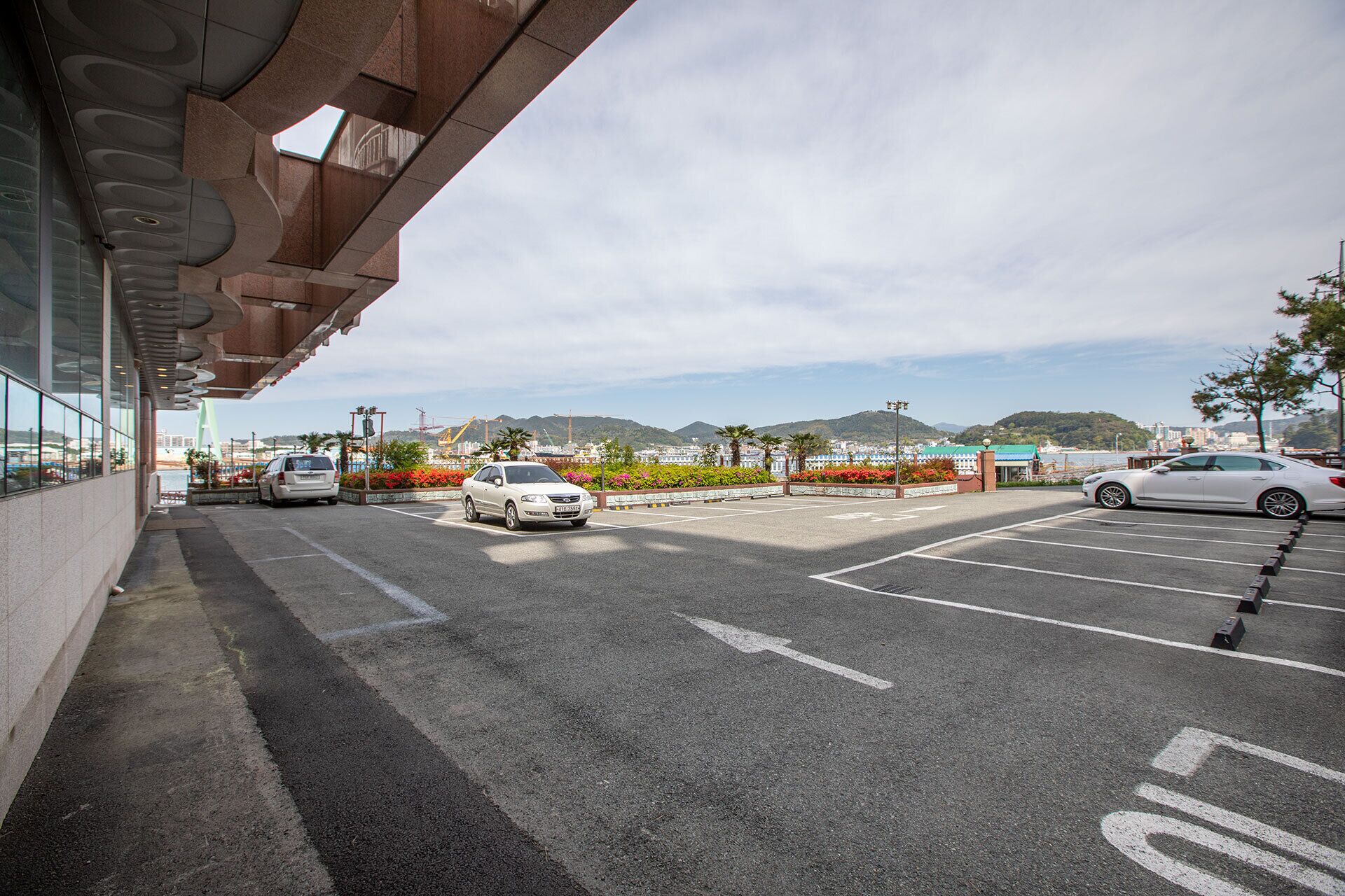 Un estacionamiento con coches, un edificio de diseño moderno y una vista de montañas y agua al fondo.