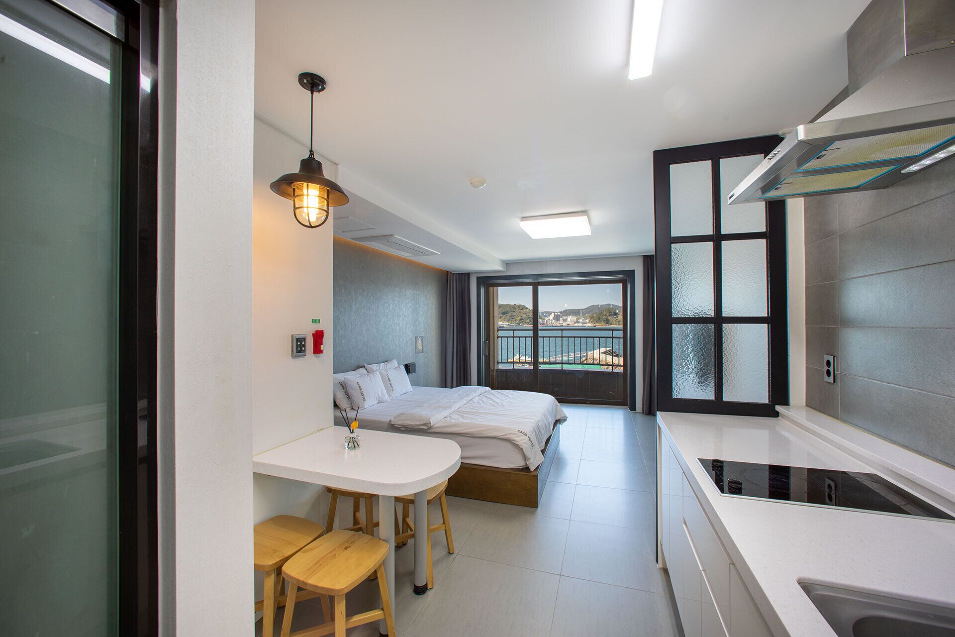Habitación de hotel moderna con una cama, un pequeño comedor con taburetes, una cocina con estufa y un ventanal con vista al mar.