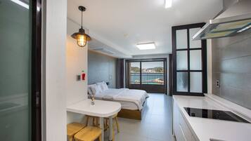 Habitación de hotel moderna con una cama, un pequeño comedor con taburetes, una cocina con estufa y un ventanal con vista al mar.