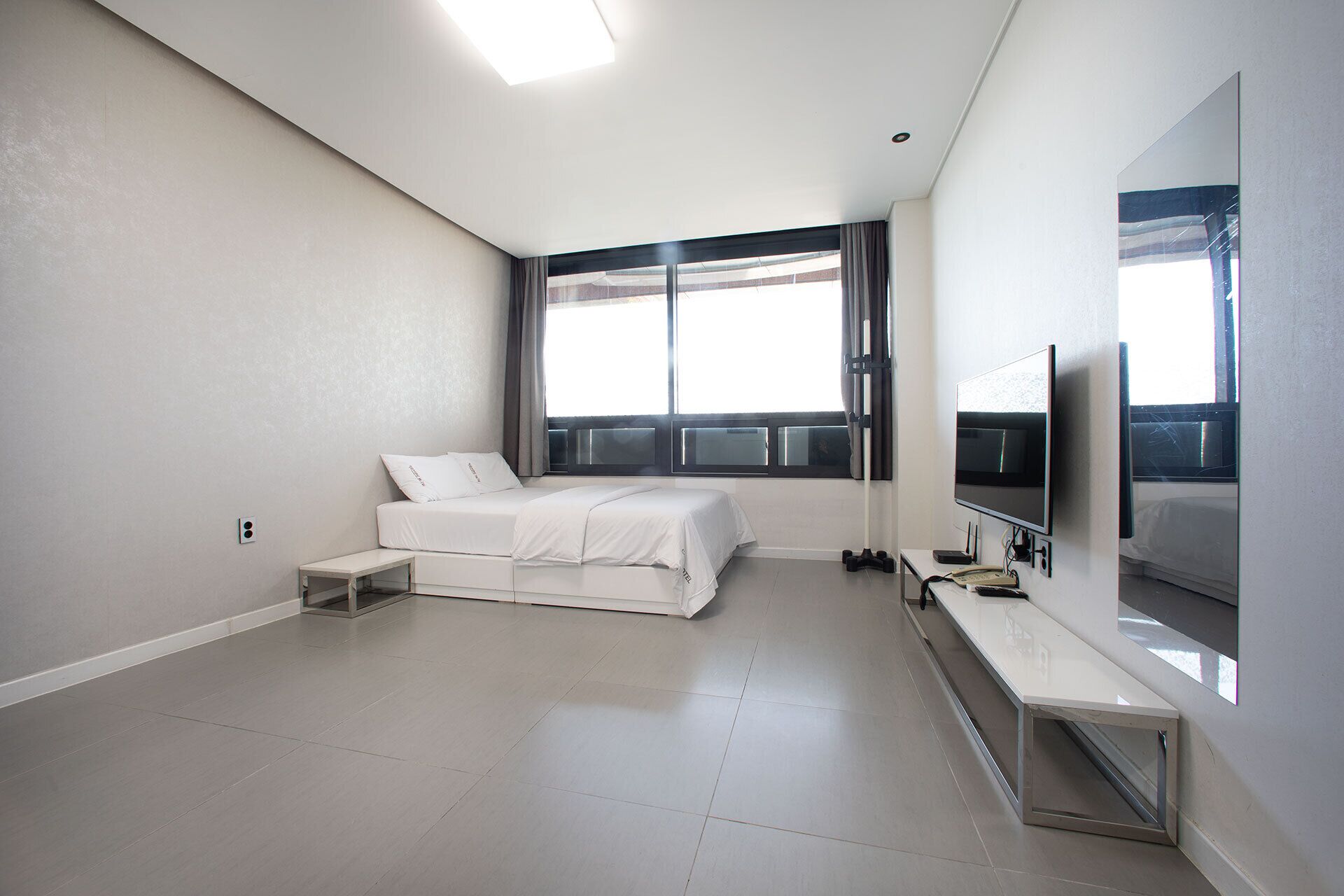 A modern hotel room with a bed, a TV, a desk, and a large window.