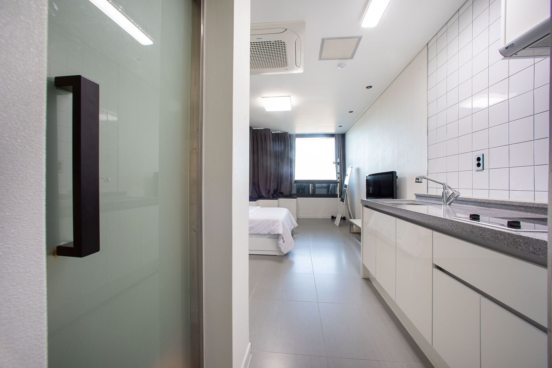 A hotel room with a kitchenette, a bed, a window with curtains, and a mirror.