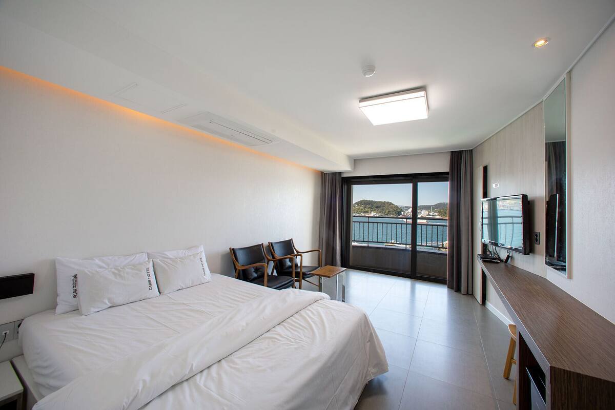 Een hotelkamer met een bed, bureau, stoel, televisie en een groot raam met uitzicht op zee.