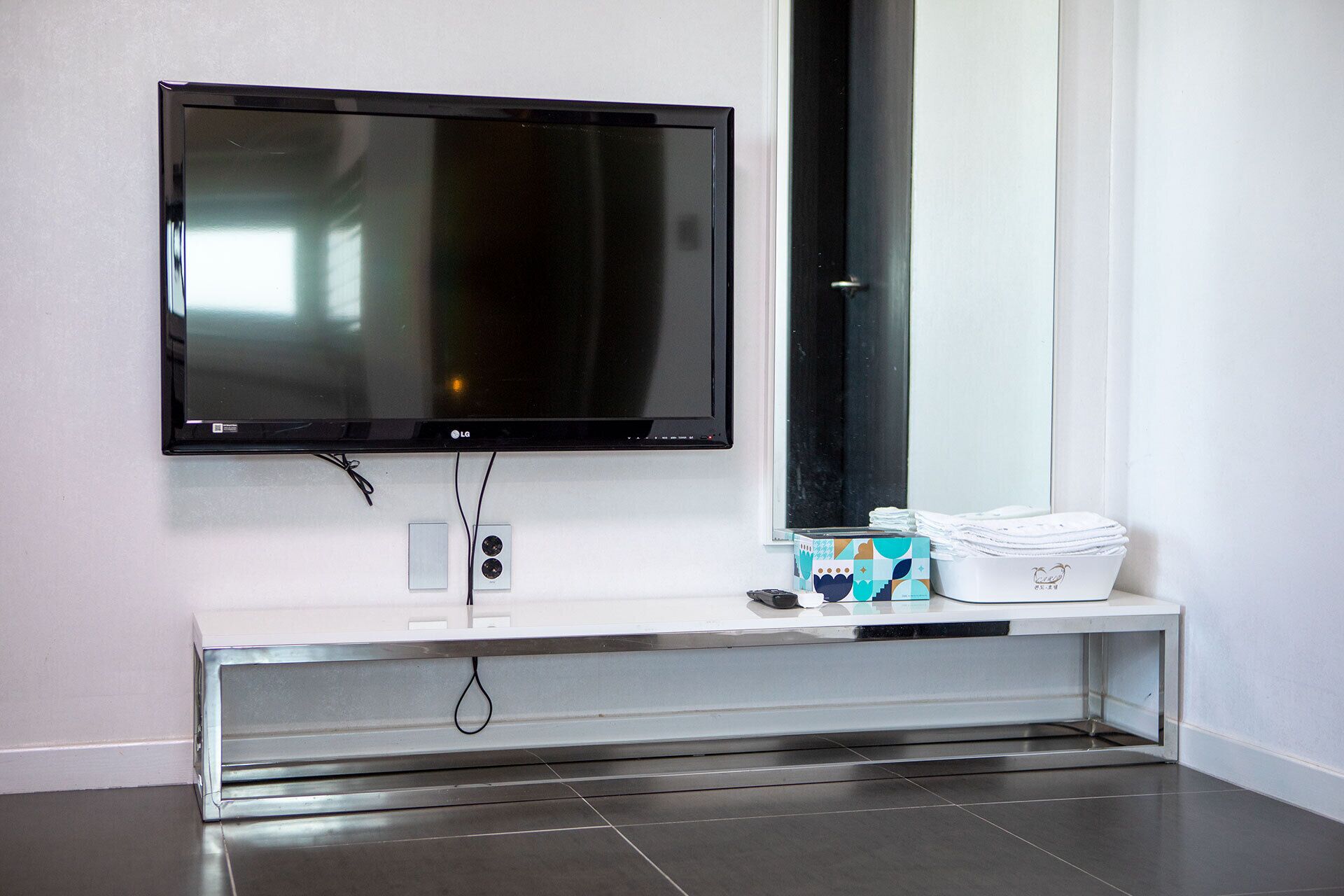 A modern bathroom with a large mirror, a flat-screen TV mounted above a stainless steel vanity, and a small shelf with toiletries.