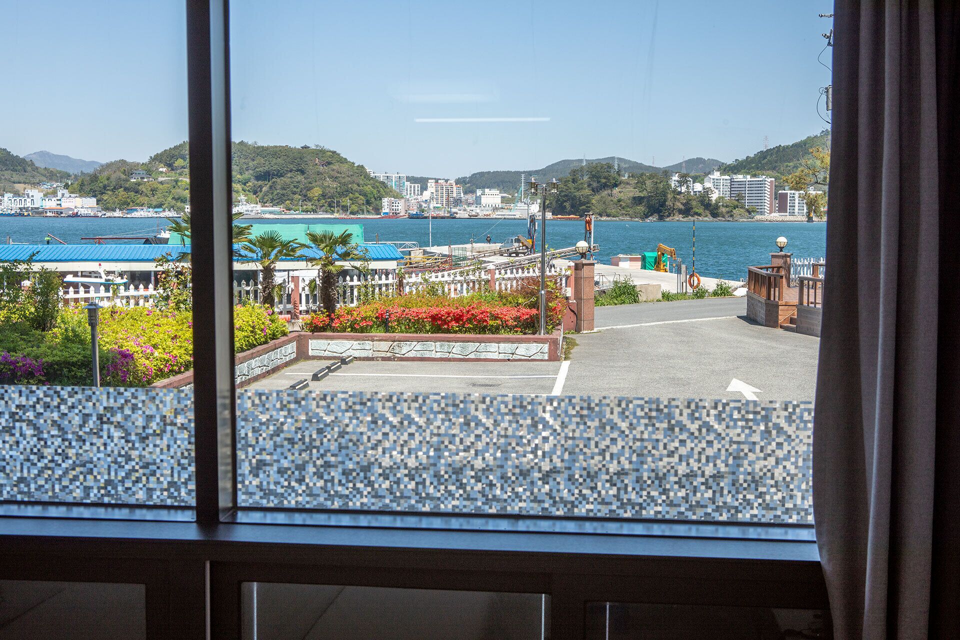 A view from a window overlooking a marina with boats, a waterfront promenade, and buildings in the background.