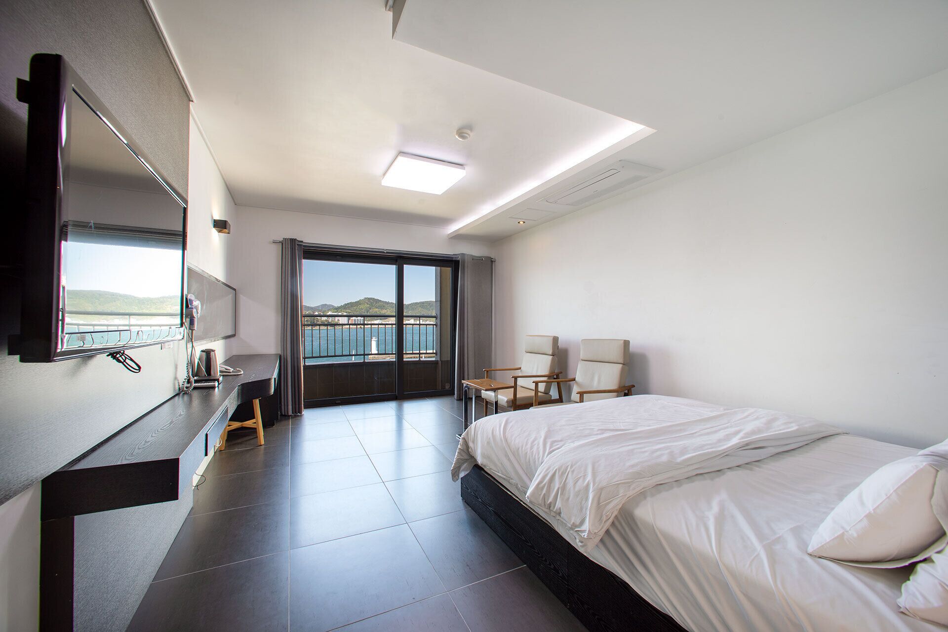 Habitación de hotel moderna con una cama grande, un escritorio, una silla y vista al mar a través de una puerta corredera.
