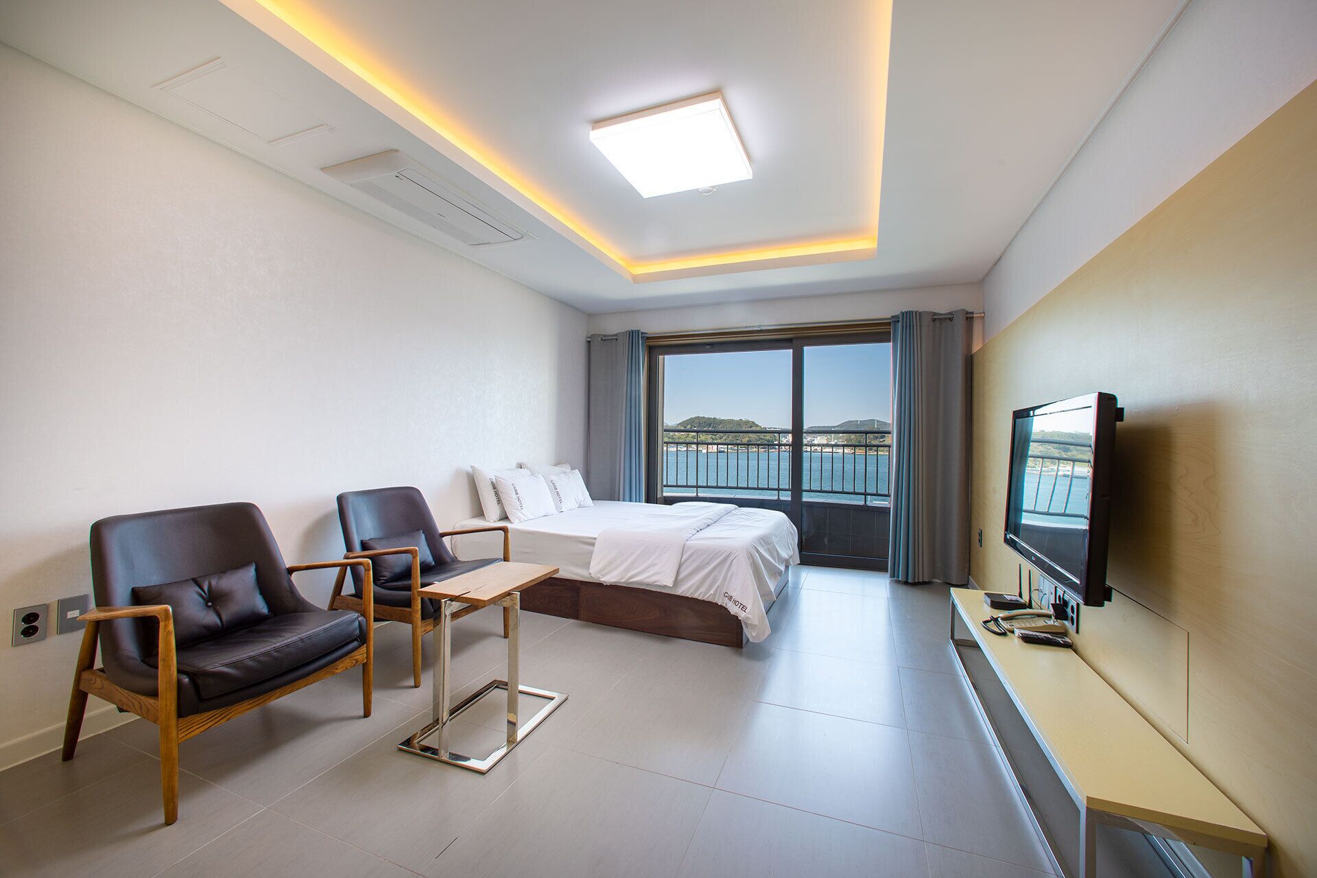 A hotel room with a bed, two chairs, a desk, a TV, and a large window with a view of the sea.