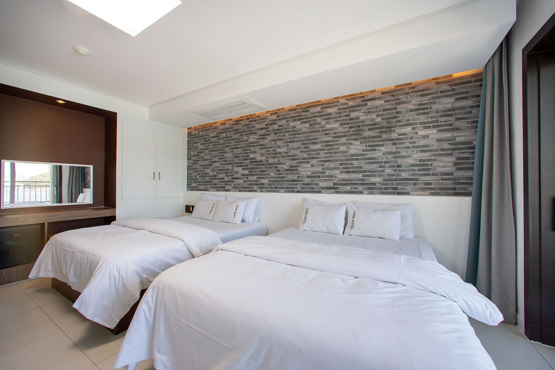 A hotel room with two beds, a large mirror, a brick-patterned wall, and a view of the outdoors.