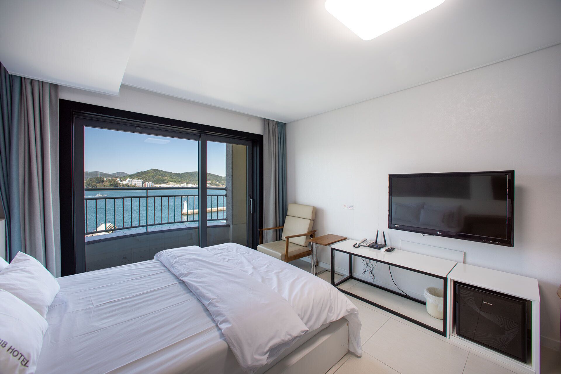 A hotel room with a bed, a TV, a desk, and a view of the sea.