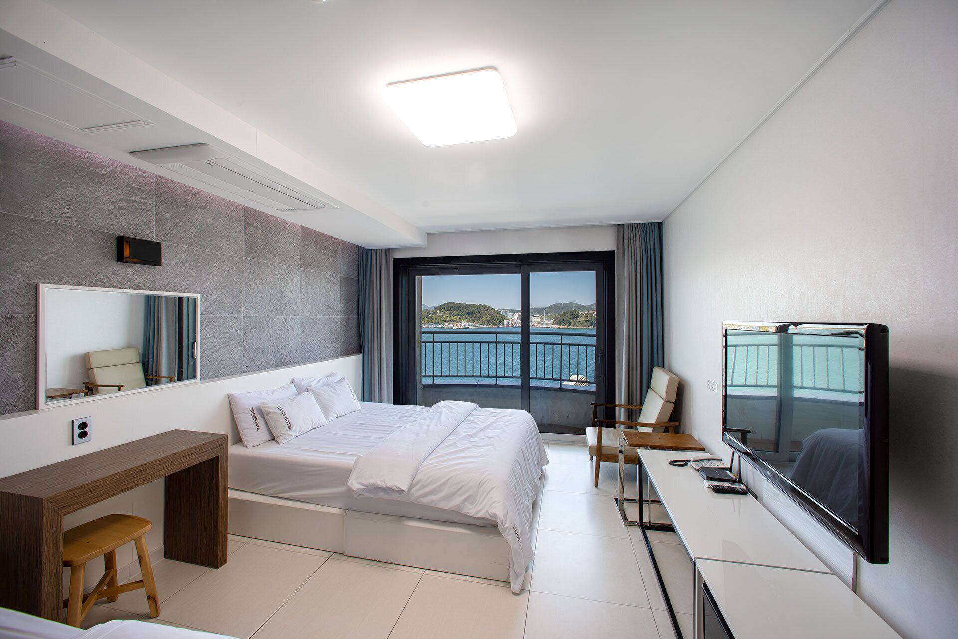 A modern hotel room with a large bed, a desk, a chair, a TV, and a view of the sea.