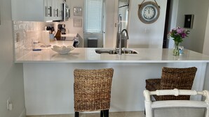 Una cocina moderna con armarios blancos, un lavamanos de acero inoxidable y una isla grande con estufa integrada. Hay sillas de mimbre y una silla blanca con cojín a rayas.
