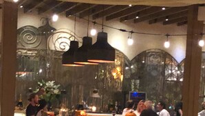 Un comedor interior con mesas y sillas de madera, un techo alto con vigas a la vista e iluminación decorativa.
