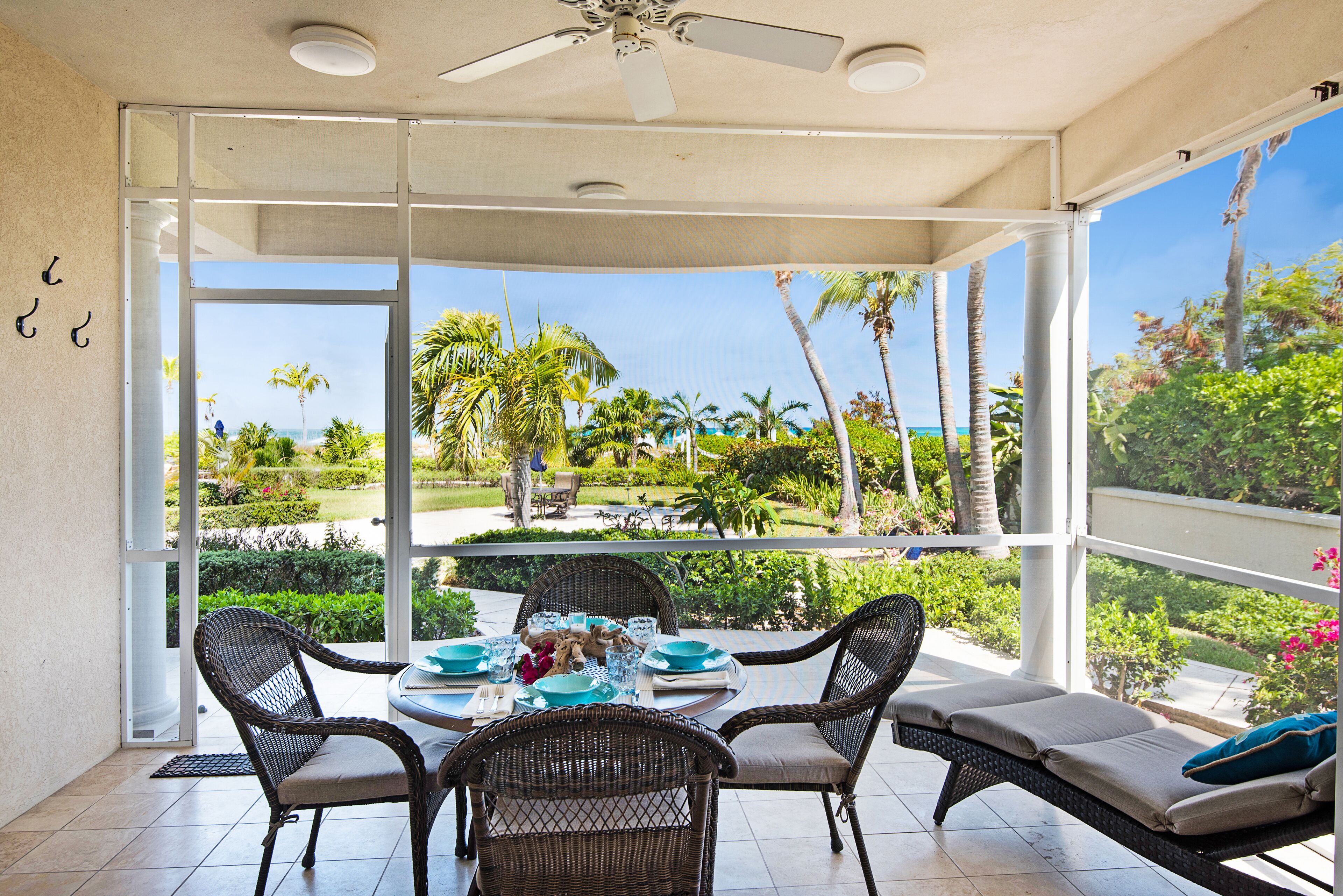 Comedor con sillas de mimbre y una mesa preparada para cuatro, con vista a un jardín tropical con palmeras y un cielo despejado.