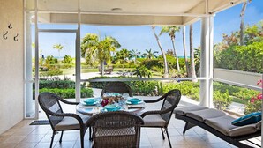 Comedor con sillas de mimbre y una mesa preparada para cuatro, con vista a un jardín tropical con palmeras y un cielo despejado.