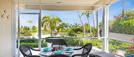 A dining area with wicker chairs and a table set for four, overlooking a tropical garden with palm trees and a clear sky.