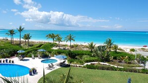 Zona de piscina con sillas de descanso y sombrillas, rodeada de palmeras y vistas a la playa y al océano.