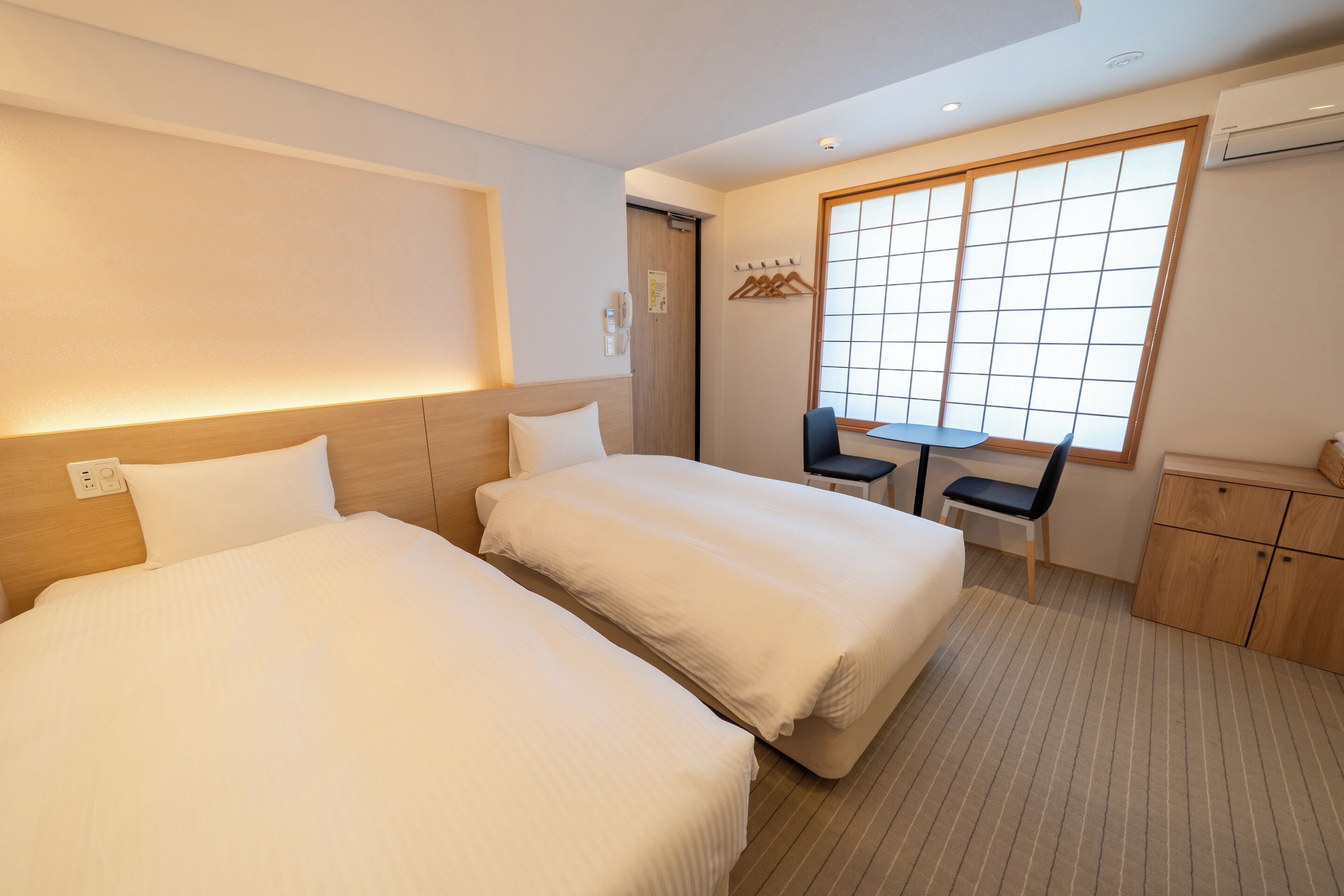 A hotel room with two beds, a small table, a chair, and a cabinet.
