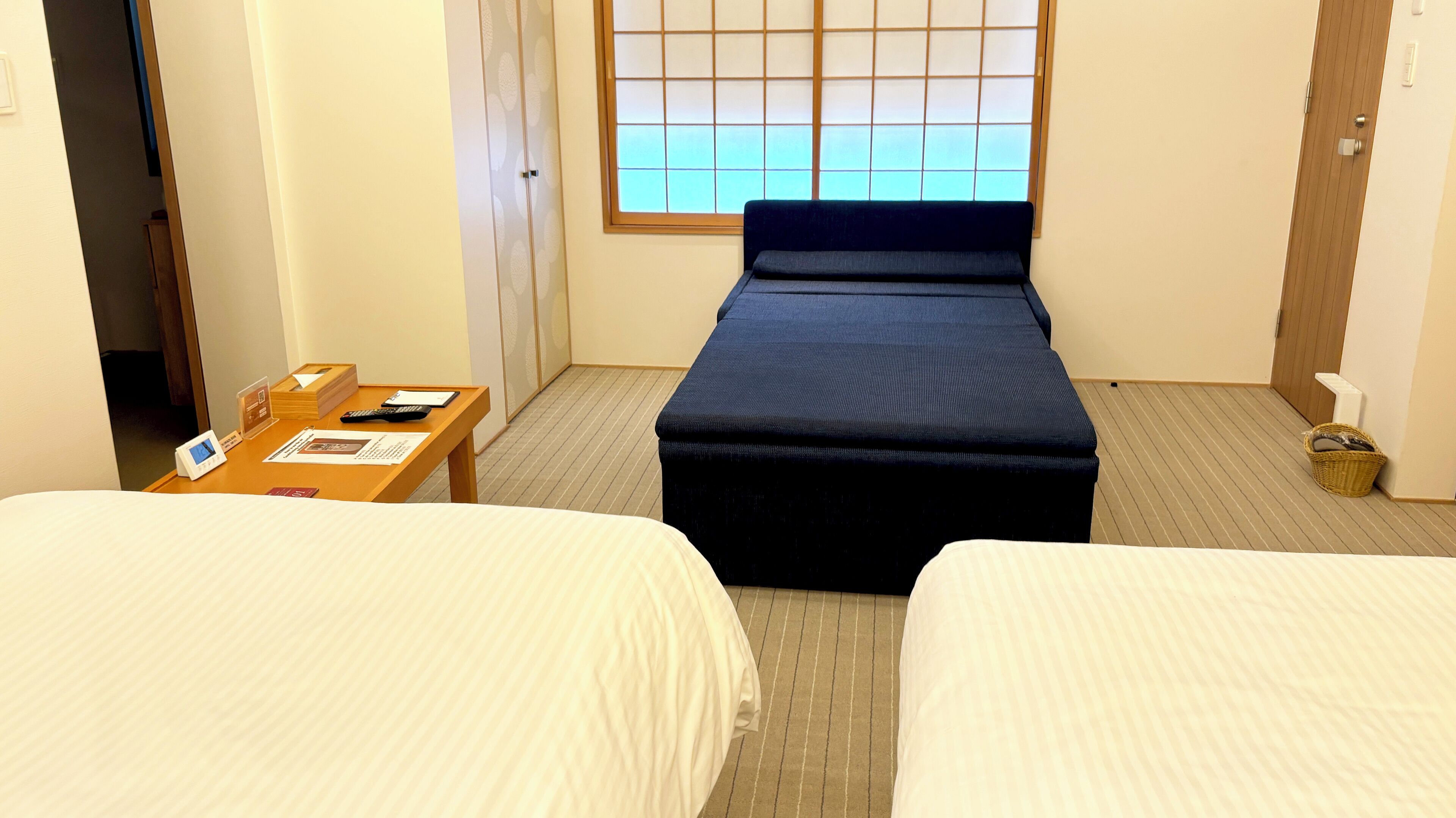 A hotel room with two beds, a wooden table with a phone and papers, a window with blinds, and a small basket on the floor.