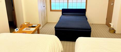 A hotel room with two beds, a wooden table with a phone and papers, a window with blinds, and a small basket on the floor.