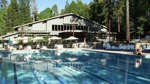 Ein Schwimmbecken mit klarem, blauen Wasser und Bahnmarkierungen, umgeben von Liegestühlen und Sonnenschirmen, mit einem Gebäude im Hintergrund.