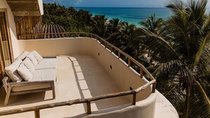 A balcony with a cushioned seating area, a railing, and a view of the ocean and palm trees.