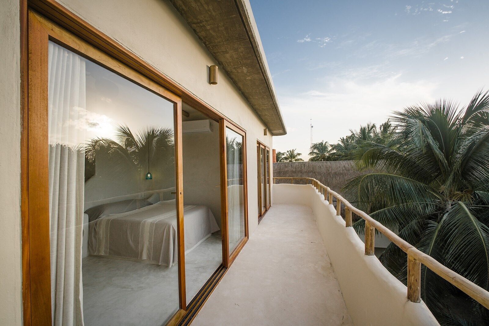 A balcony with a view of palm trees, a bed with white bedding, and a concrete wall.