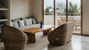 A spacious living area with a wooden ceiling, a large window offering a view of palm trees, and a cozy seating arrangement with a sofa and two wicker chairs.
